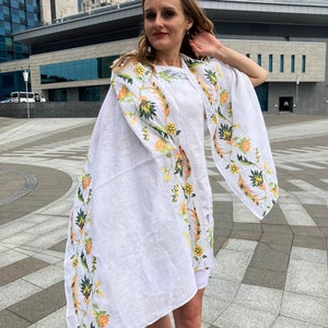 May include: A white linen shawl with a floral embroidery design in shades of yellow, green, and orange. The shawl is draped over a person's shoulders and is flowing in the wind.
