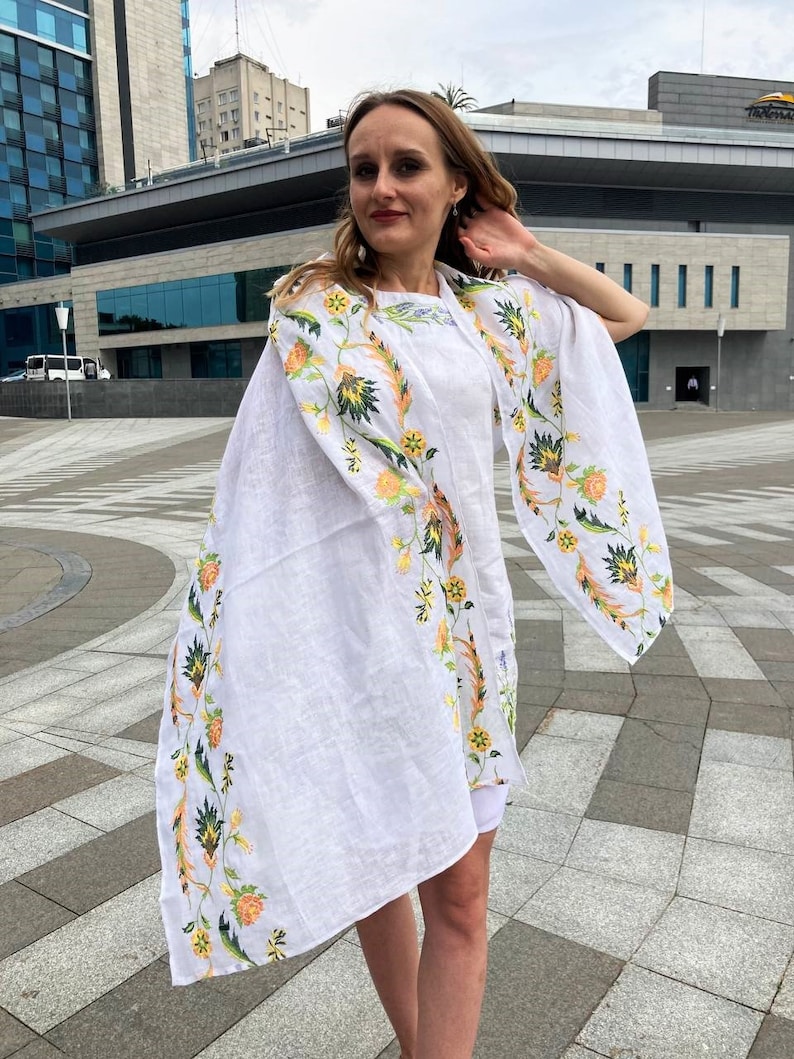 May include: A white linen shawl with a floral embroidery design in shades of yellow, green, and orange. The shawl is draped over a person's shoulders and is flowing in the wind.