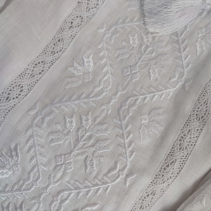 Peut inclure: Tissu de lin blanc avec un d&eacute;licat motif brod&eacute; blanc pr&eacute;sentant des formes g&eacute;om&eacute;triques et des motifs floraux. Le tissu est bord&eacute; d'une dentelle.