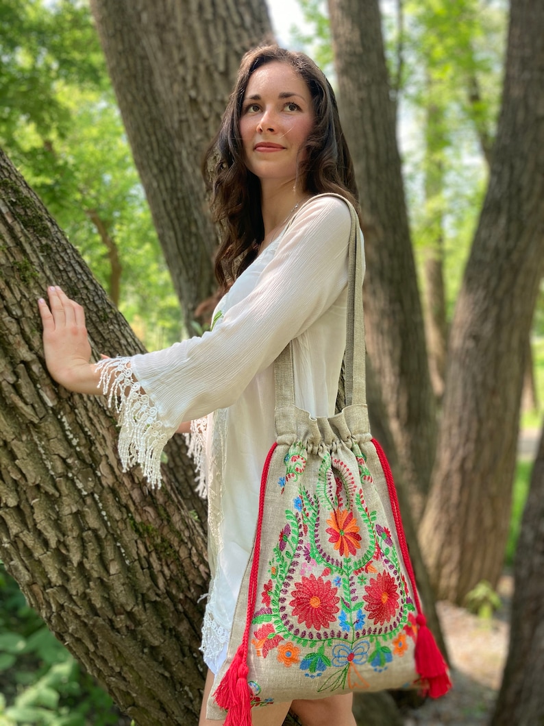 May include: A beige linen tote bag with red, green, blue, and orange floral embroidery. The bag has red tassel drawstrings.