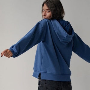May include: A woman wearing a blue hooded sweatshirt with a relaxed fit. The sweatshirt has a side seam detail and is made of a soft, comfortable fabric.