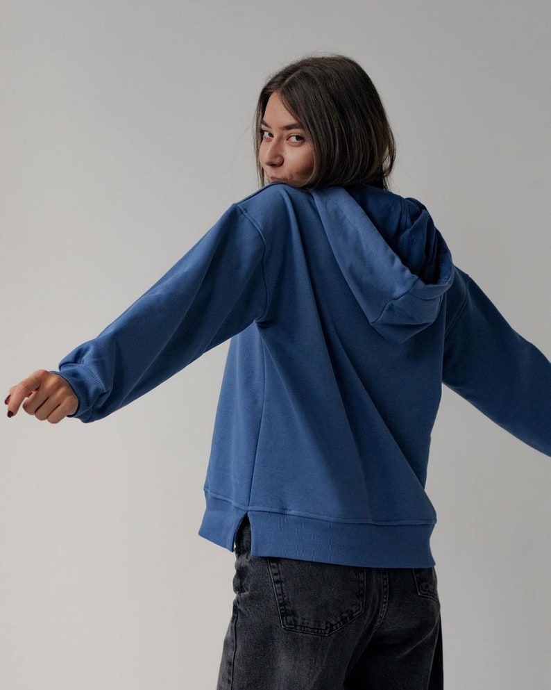 May include: A woman wearing a blue hooded sweatshirt with a relaxed fit. The sweatshirt has a side seam detail and is made of a soft, comfortable fabric.