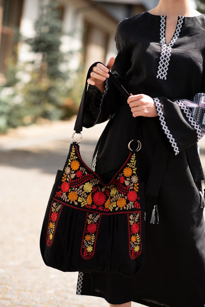 Puede incluir: Un bolso tote negro con una correa larga y bordado floral de colores. El bolso tiene un detalle fruncido en la parte superior y un borla en el lateral.
