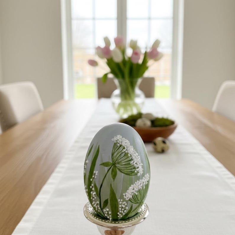 K&ouml;nnte beinhalten: Ein handbemaltes Osterei mit floralem Design. Das Ei ist hellblau mit gr&uuml;nen Bl&auml;ttern und wei&szlig;en Bl&uuml;ten. Es steht in einem silbernen Eierbecher auf einem wei&szlig;en Tischl&auml;ufer. Im Hintergrund steht eine Vase mit rosa und wei&szlig;en Tulpen.