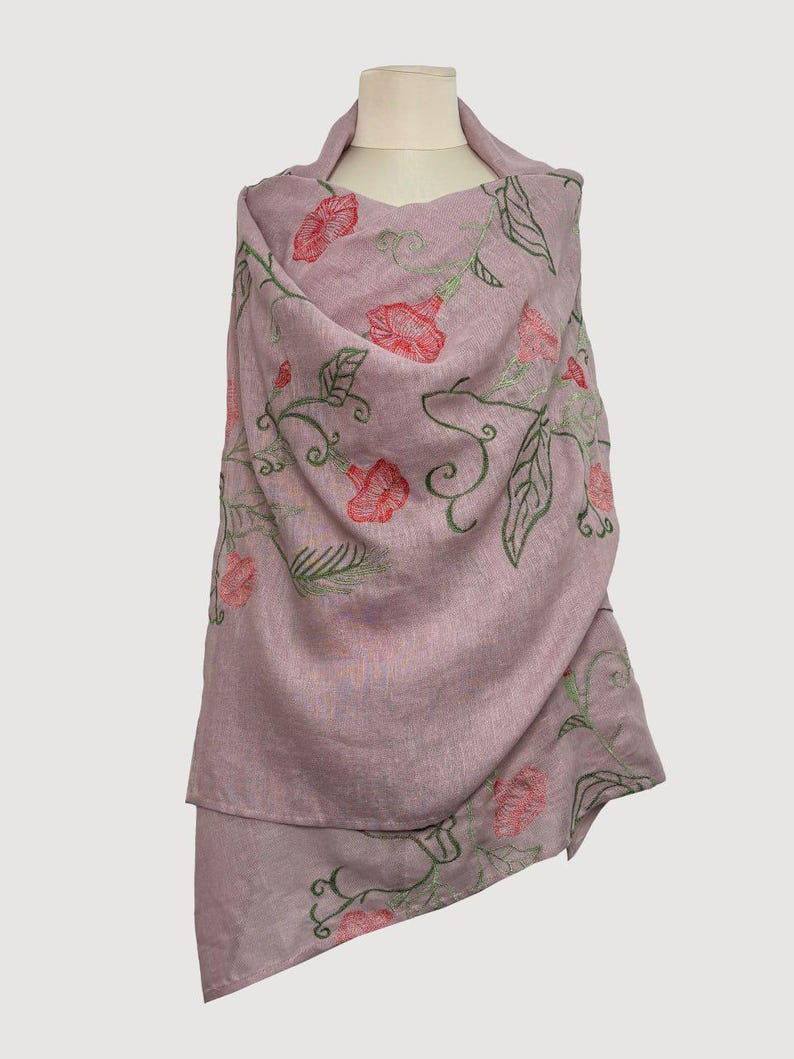 May include: A mauve shawl featuring embroidered red flowers and green leaves. The floral pattern adorns the shawl, displayed on a mannequin. The shawl is made from a soft, flowing material.