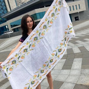 May include: A white linen tablecloth with a floral embroidery design in shades of yellow, orange, and green. The embroidery features stylized flowers and leaves.