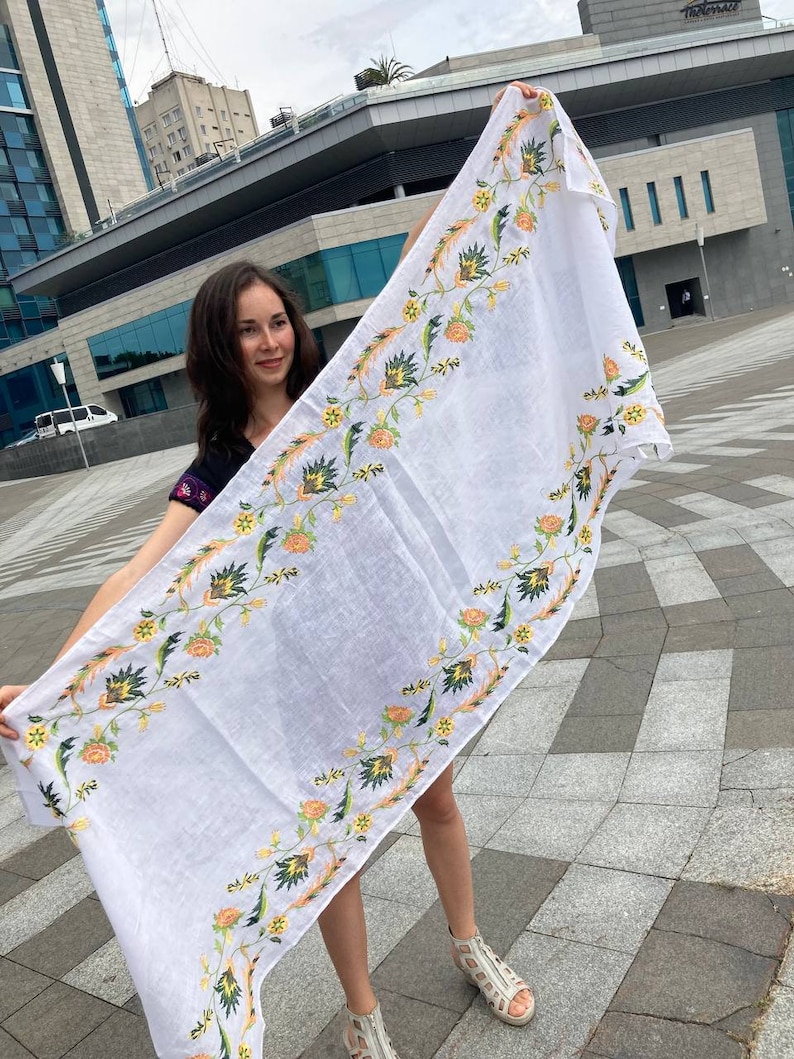 May include: A white linen tablecloth with a floral embroidery design in shades of yellow, orange, and green. The embroidery features stylized flowers and leaves.