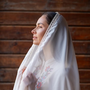Puede incluir: Una mujer con un velo blanco y una camisa blanca bordada. El velo tiene un ribete de encaje decorativo. La camisa presenta bordados florales en rojo, azul y verde. El fondo es una pared de madera.