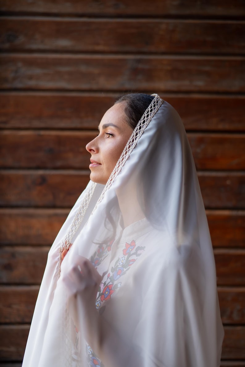Puede incluir: Una mujer con un velo blanco y una camisa blanca bordada. El velo tiene un ribete de encaje decorativo. La camisa presenta bordados florales en rojo, azul y verde. El fondo es una pared de madera.