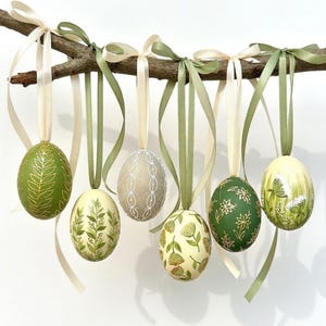 Puede incluir: Un conjunto de huevos de Pascua decorativos colgados de una rama. Cada huevo está pintado con diseños florales y de hojas en tonos verdes, crema y dorado. Los huevos están suspendidos por cintas crema y verdes, creando una exhibición festiva.