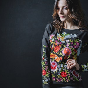 May include: Black long-sleeved sweatshirt with colourful floral embroidery. The design features flowers, birds, and foliage in red, orange, pink, green, and blue. The embroidery is detailed and covers the front and sleeves of the sweatshirt.