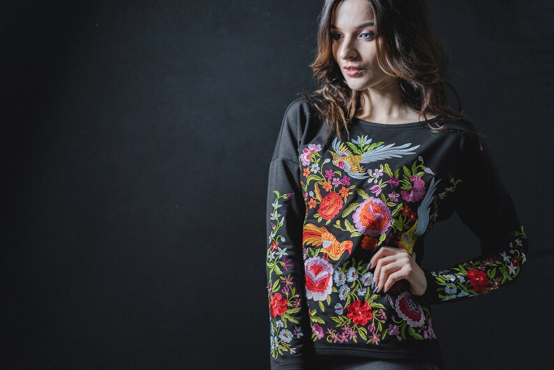 May include: Black long-sleeved sweatshirt with colourful floral embroidery. The design features flowers, birds, and foliage in red, orange, pink, green, and blue. The embroidery is detailed and covers the front and sleeves of the sweatshirt.