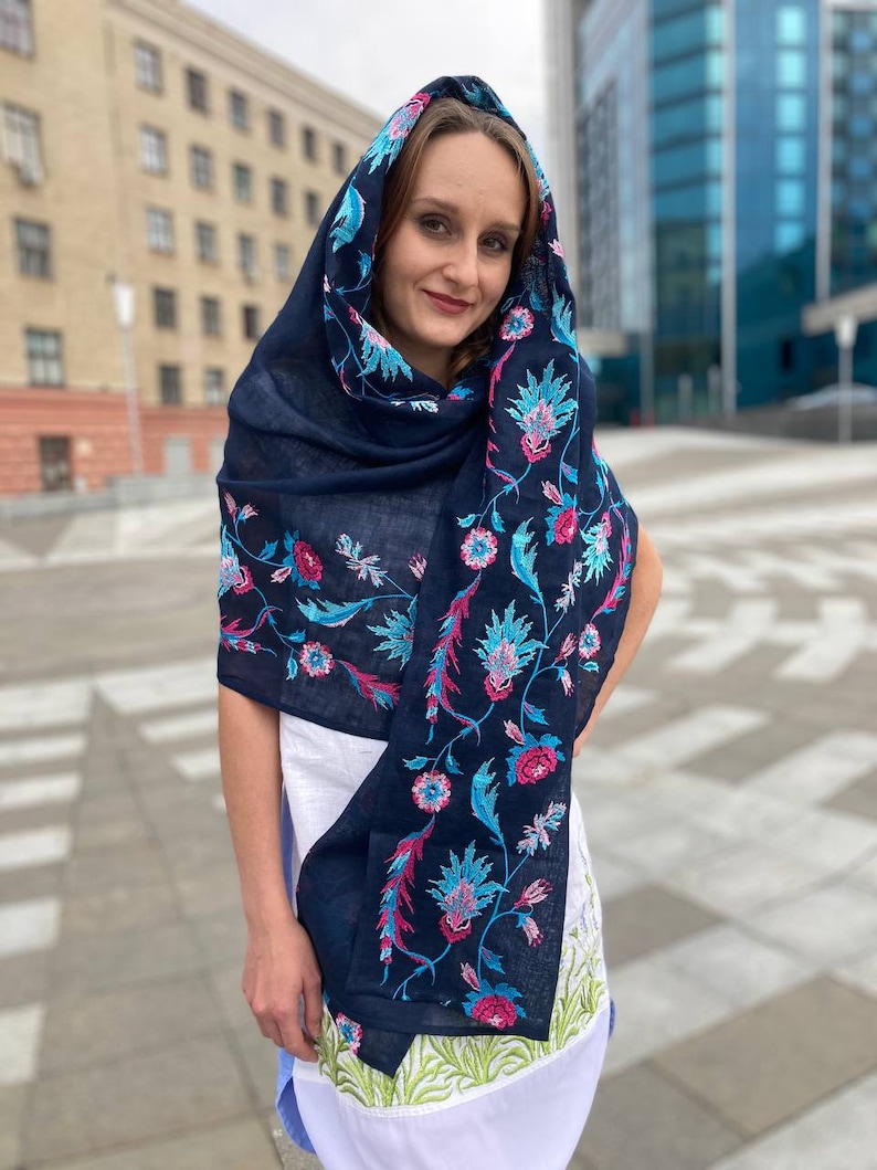 May include: A navy blue scarf with floral embroidery in pink, blue, and green. The scarf is draped over a person's head and shoulders.