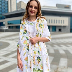 May include: A white linen shawl with a floral pattern in shades of green, yellow, and orange. The shawl is draped over a person's shoulders and is tied at the neck.