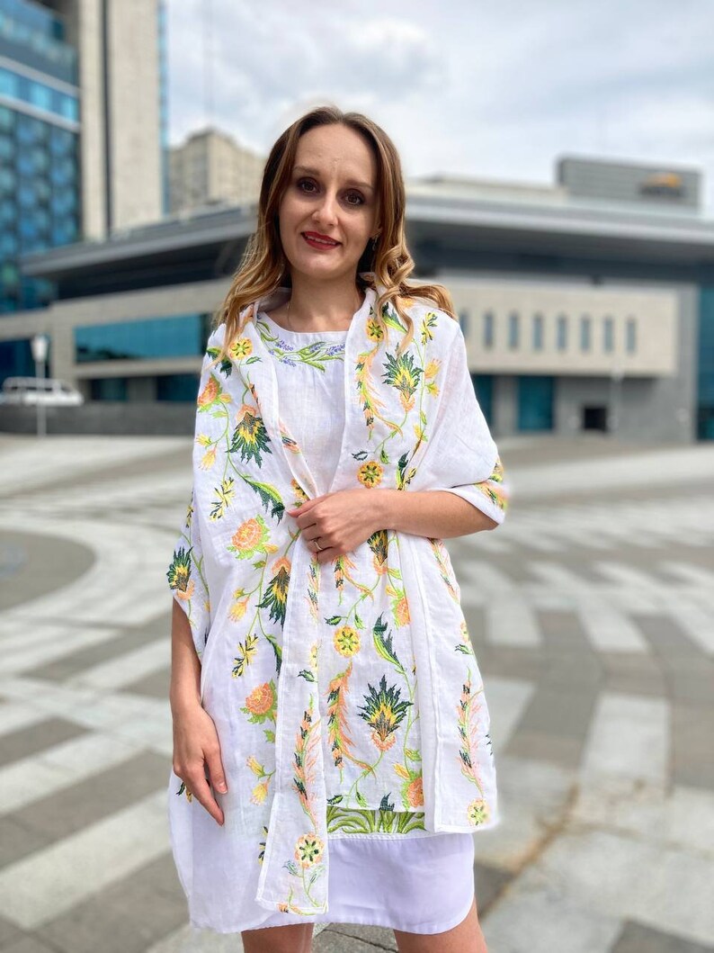 May include: A white linen shawl with a floral pattern in shades of green, yellow, and orange. The shawl is draped over a person's shoulders and is tied at the neck.