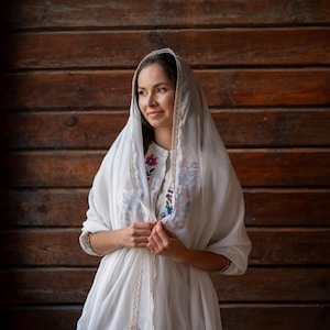 Puede incluir: Una mujer con un vestido blanco y un velo, adornada con delicados bordes de encaje. El vestido presenta bordados florales coloridos en el corpi&ntilde;o. El fondo es una pared de madera.
