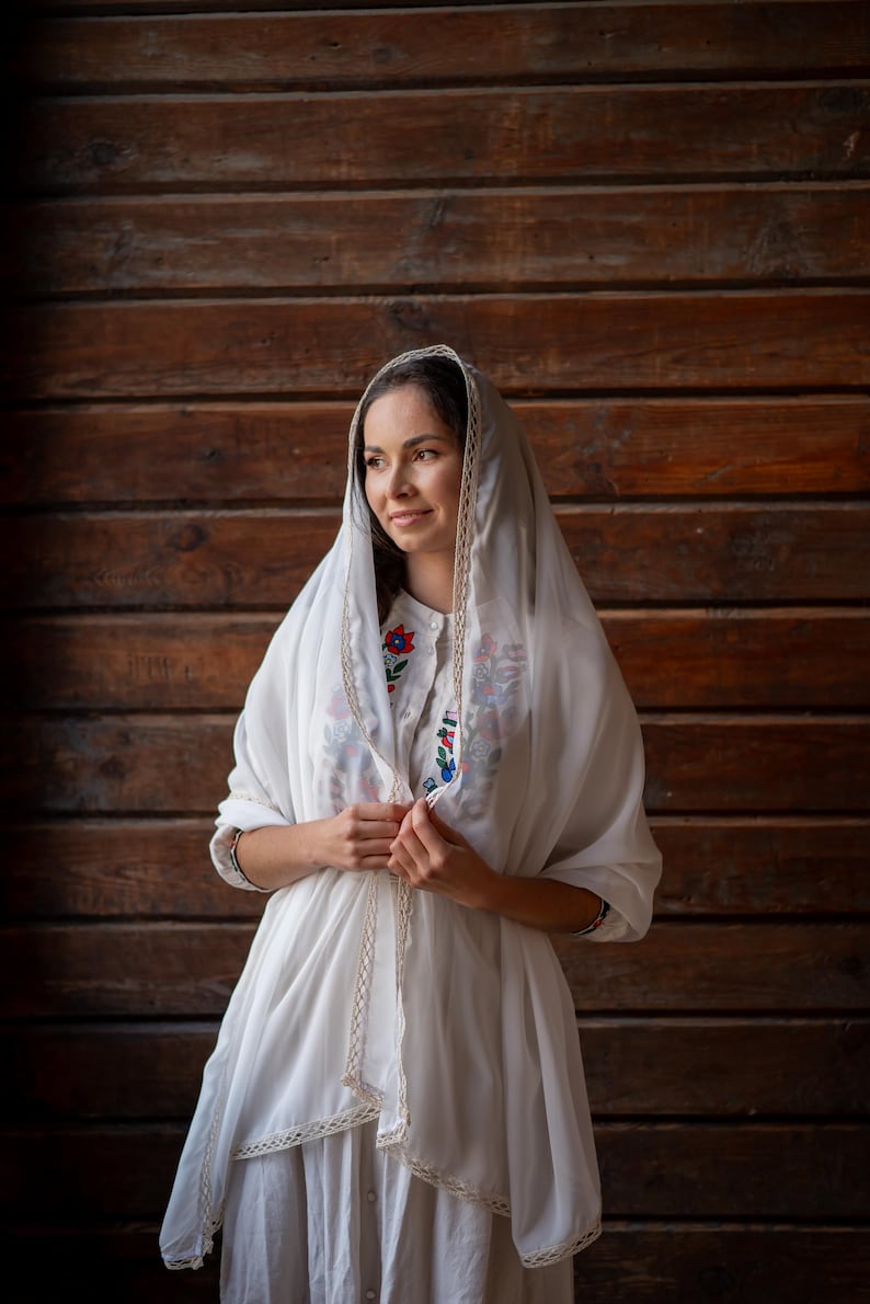Puede incluir: Una mujer con un vestido blanco y un velo, adornada con delicados bordes de encaje. El vestido presenta bordados florales coloridos en el corpi&ntilde;o. El fondo es una pared de madera.