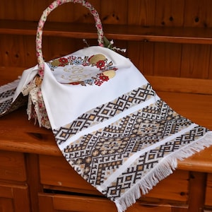 May include: A white embroidered cloth with a geometric pattern in brown, gold, and white, fringed at the edge, draped over a wooden surface. The cloth is part of a basket with a floral handle and lining, and floral embroidery.