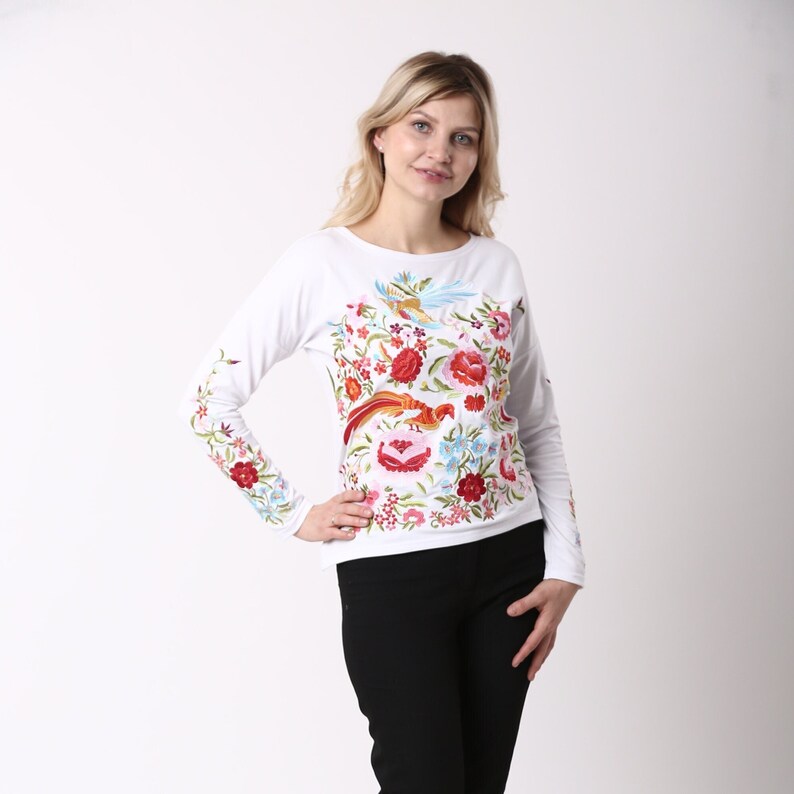 May include: White long-sleeved shirt with vibrant floral and bird embroidery. The design includes red, pink, blue, and green flowers and birds. The embroidery extends to the sleeves. The shirt is worn with black trousers.