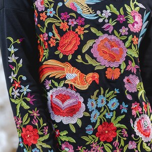 May include: Black long-sleeved top with colourful floral and bird embroidery. The design includes red, orange, pink, blue, and purple flowers, and two embroidered birds. The detailed embroidery covers the front and sleeves.