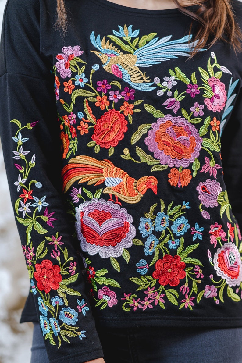 May include: Black long-sleeved top with colourful floral and bird embroidery. The design includes red, orange, pink, blue, and purple flowers, and two embroidered birds. The detailed embroidery covers the front and sleeves.