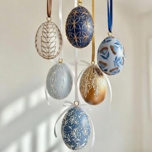 May include: Six decorative Easter eggs suspended by ribbons. The eggs are painted in various colours, including white, blue, and gold, with intricate floral and geometric patterns. Each egg is suspended by a ribbon, creating a festive display.