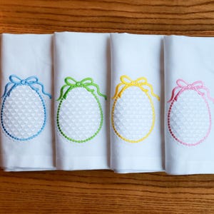 Può includere: Quattro tovaglioli di stoffa bianchi con ricami a forma di uovo. Ogni uovo è delineato con un colore diverso: blu, verde, giallo e rosa. Le uova hanno un motivo strutturato e un fiocco in alto.