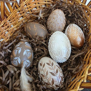 Könnte beinhalten: Ein Weidenkorb gefüllt mit dekorativen Ostereiern. Die Eier sind in verschiedenen Farben bemalt, darunter Braun, Gold und Weiß, und mit floralen und geometrischen Mustern verziert. Der Korb ist mit geschreddertem Papier und einer Feder ausgekleidet.