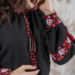 May include: A black embroidered blouse with red and white floral patterns. The blouse has long sleeves with a tassel detail at the neckline.