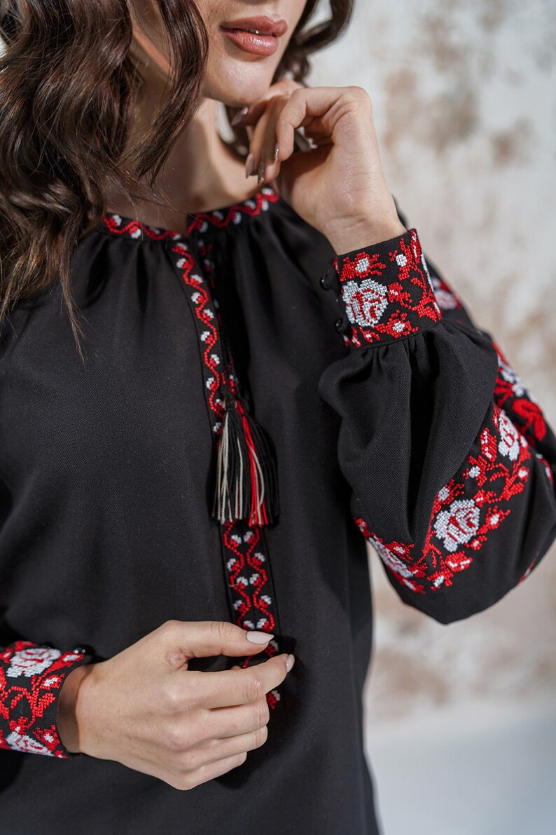 May include: A black embroidered blouse with red and white floral patterns. The blouse has long sleeves with a tassel detail at the neckline.
