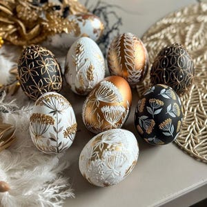 May include: A selection of decorated Easter eggs in white, black, and gold. The eggs are adorned with detailed floral and geometric patterns. Some have a metallic gold finish, others are matte. The overall style is elegant and celebratory, perfect for Easter.