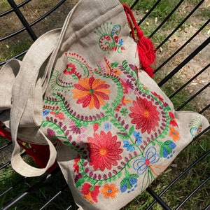 May include: A beige linen tote bag with a red tassel. The bag is embroidered with colorful flowers and vines.