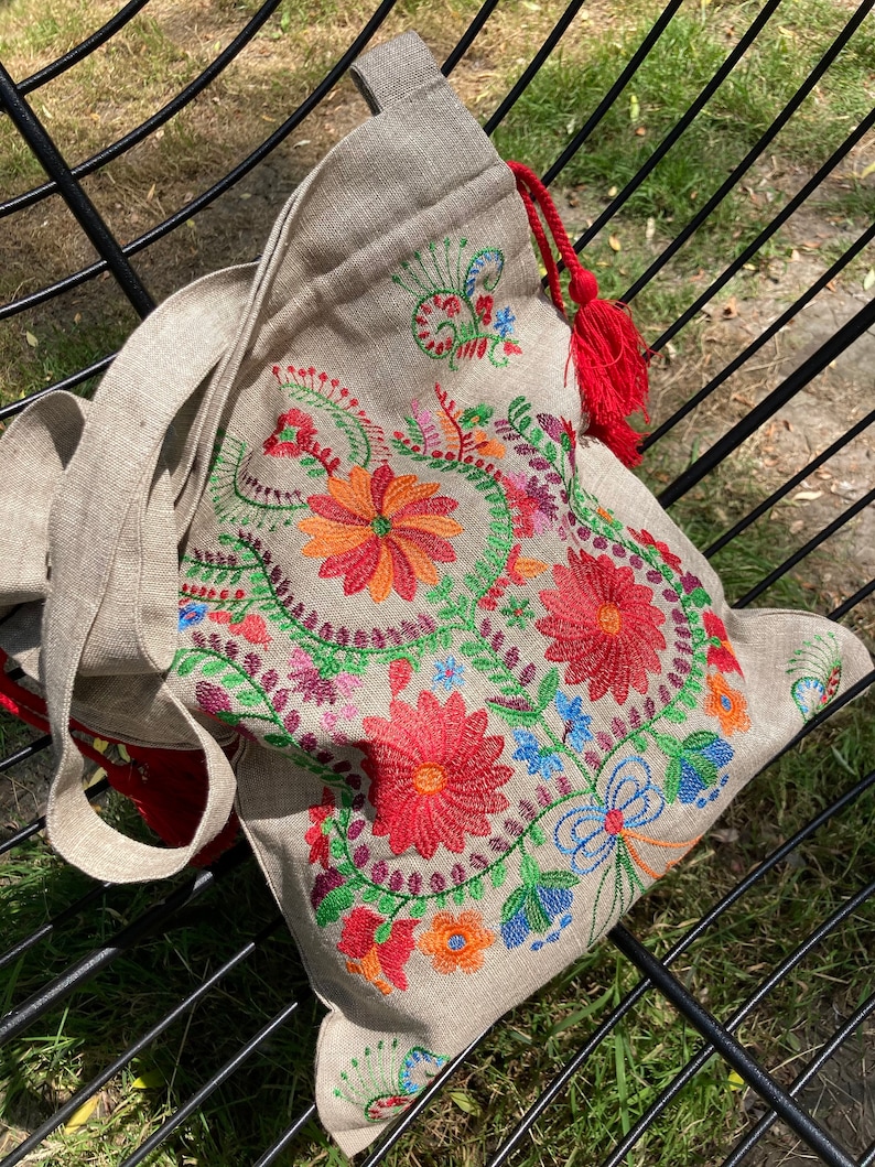 May include: A beige linen tote bag with a red tassel. The bag is embroidered with colorful flowers and vines.