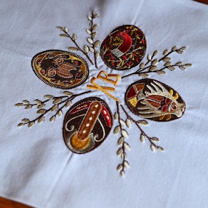 May include: Embroidered Easter egg design on a light blue fabric. The design features five decorated eggs in brown, red, and gold, arranged around sprigs of pussy willow. The letters "XP" are embroidered in gold.