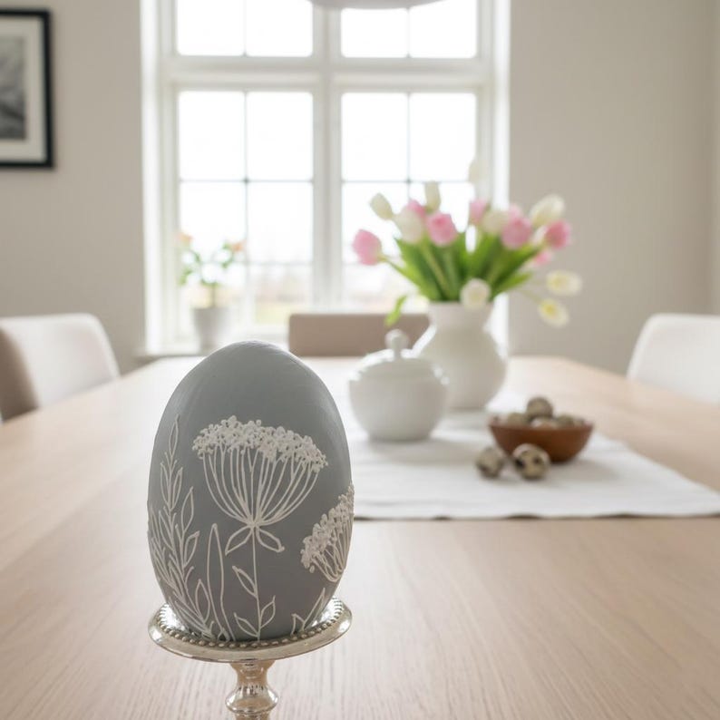 K&ouml;nnte beinhalten: Ein graues Deko-Ei mit wei&szlig;en Blumenmustern, pr&auml;sentiert auf einem silbernen St&auml;nder. Das Ei ist der Mittelpunkt, vor dem Hintergrund eines Esstisches mit einer Vase mit rosa und wei&szlig;en Tulpen.