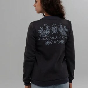 Puede incluir: Una sudadera gris oscuro con un dise&ntilde;o bordado blanco en la espalda. El dise&ntilde;o presenta tres p&aacute;jaros estilizados y patrones geom&eacute;tricos.