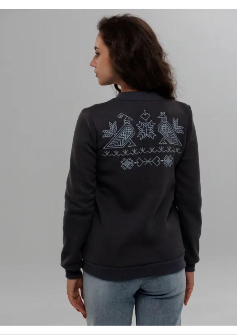 Puede incluir: Una sudadera gris oscuro con un dise&ntilde;o bordado blanco en la espalda. El dise&ntilde;o presenta tres p&aacute;jaros estilizados y patrones geom&eacute;tricos.