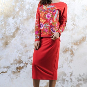 May include: A red two-piece outfit comprising a long-sleeved top and a knee-length skirt. The top is decorated with vibrant floral embroidery. The outfit is worn with platform high heels.