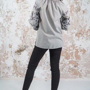 May include: A gray blouse with black embroidered floral designs on the sleeves. The blouse has a gathered neckline and long sleeves. The model is wearing black pants and high heels.