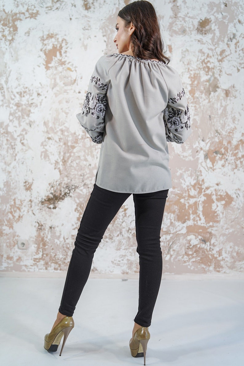 May include: A gray blouse with black embroidered floral designs on the sleeves. The blouse has a gathered neckline and long sleeves. The model is wearing black pants and high heels.