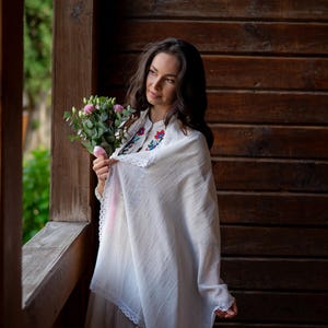 Puede incluir: Una mujer con un vestido y chal blancos, sosteniendo un ramo de flores rosas. El chal tiene un ribete de encaje y detalles florales bordados. El fondo presenta una pared de madera y vegetaci&oacute;n.