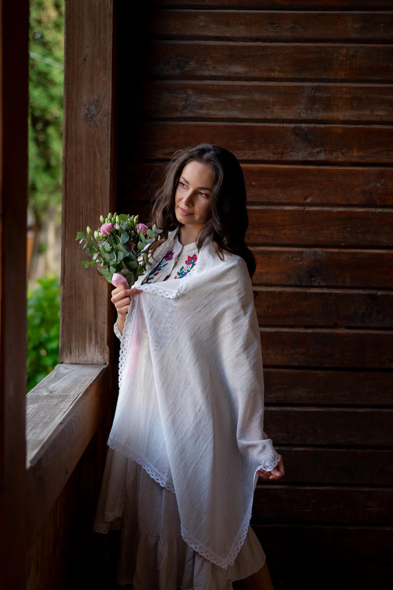 Puede incluir: Una mujer con un vestido y chal blancos, sosteniendo un ramo de flores rosas. El chal tiene un ribete de encaje y detalles florales bordados. El fondo presenta una pared de madera y vegetaci&oacute;n.