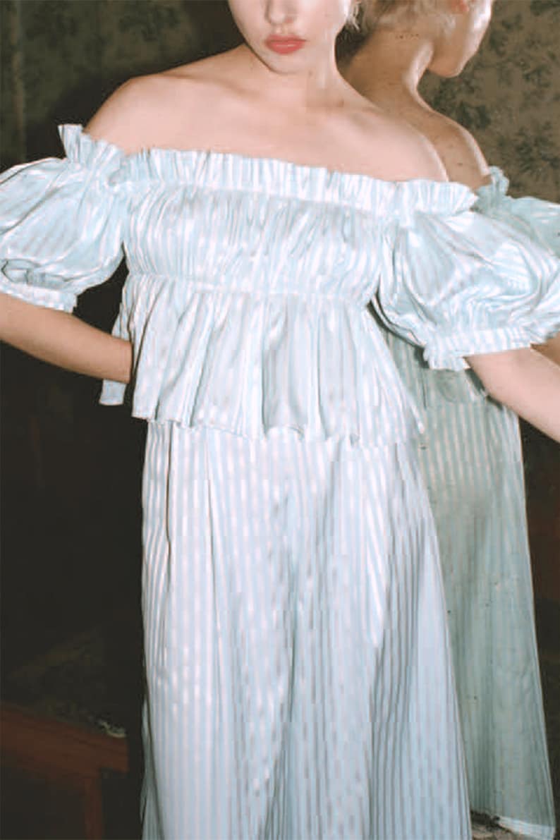 Puede incluir: Una blusa azul claro de hombros descubiertos con rayas blancas y un escote con volantes. La blusa est&aacute; hecha de una tela sedosa y tiene mangas cortas y abullonadas.