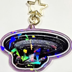 May include: A purple and holographic keychain with a spaceship design. The keychain has a gold-coloured star-shaped clasp and a circular ring. The spaceship design is detailed with various colours and shapes.