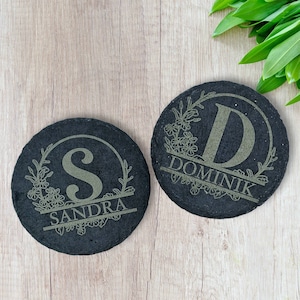 Könnte beinhalten: Zwei schwarze Schieferuntersetzer mit einem floralen Kranzdesign und einem Buchstaben in der Mitte. Die Untersetzer sind mit den Namen "Sandra" und "Dominik" personalisiert.