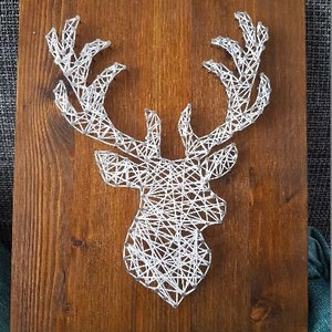 Könnte beinhalten: Ein weißer String-Art-Hirschkopf auf braunem Holzgrund.