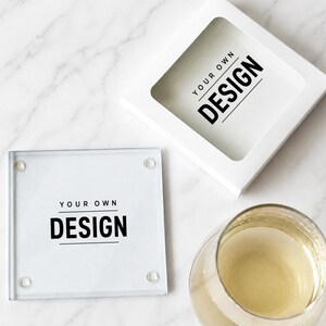May include: A square glass coaster with four small clear rubber feet and the black text YOUR OWN DESIGN printed in the center, accompanied by a matching white square gift box with a window and a glass of light-colored liquid nearby.