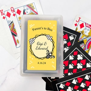 May include: A deck of playing cards in a clear plastic case. The card design features a yellow background with the text "Parent's to Bee," the names "Gigi & Edward," and the date 4.16.24, along with a bee and honeycomb design. Other playing cards are scattered around.