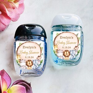 May include: Two small bottles of hand sanitizer with labels featuring a woodland animal theme. The labels are decorated with a woodgrain background, green leaves, and a variety of animals including a giraffe, lion, elephant, and monkey. The text on the labels reads "Evelyn's Baby Shower 6.19.24".