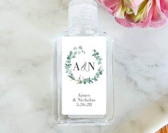 May include: Clear hand sanitizer bottle with a white label featuring a green eucalyptus wreath around the letters "A&N". Below the wreath, the label reads "Aimee & Nicholas 5.26.28". The bottle is on a marble surface.
